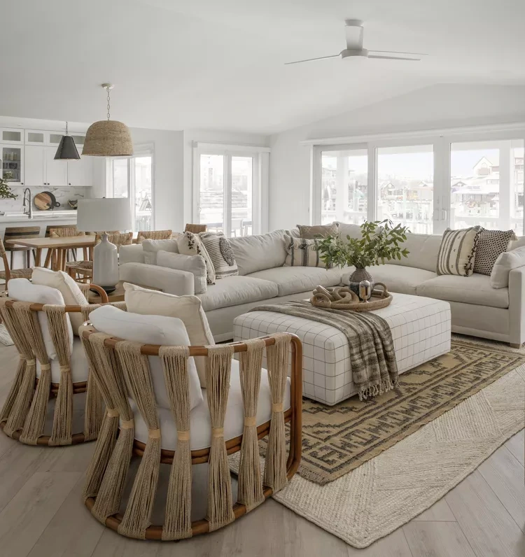 Sala de estar com decoração boho neutra, sofá em L claro, poltronas de madeira com amarrações em corda, mesa de centro estofada quadriculada, tapetes sobrepostos e janelas amplas que iluminam todo o ambiente.
