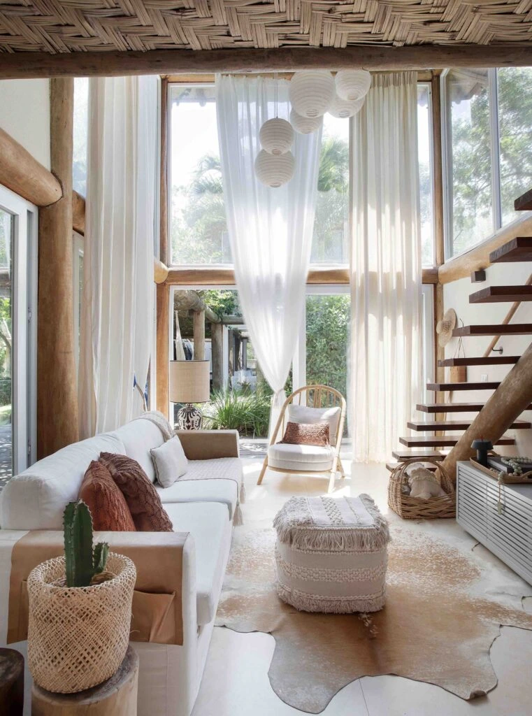 Sala de estar em estilo Boho Chic com sofá branco, almofadas em tons terrosos, pufe com franjas, tapete de couro e teto de palha trançada. Grandes janelas deixam a luz natural entrar, destacando a madeira rústica e a leveza das cortinas brancas.