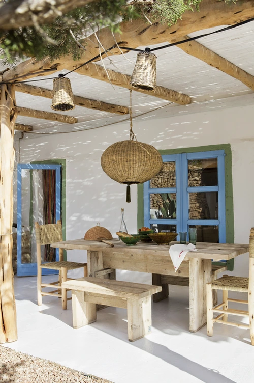 Varanda com mesa e bancos de madeira rústica, cadeiras de palha, luminária artesanal pendente e molduras de janelas em azul e verde, em ambiente com cobertura de madeira.