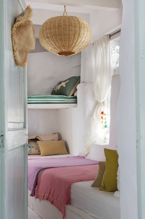 Quarto boho compacto com beliches embutidos, roupa de cama rosa e verde, almofadas coloridas e luminária pendente de palha, com luz natural entrando pela janela com cortinas leves.