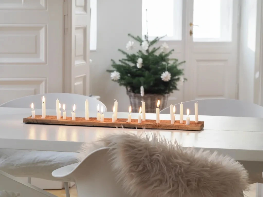 Mesa de jantar no estilo escandinavo com velas minimalistas sobre suporte de madeira, árvore natural ao fundo e cadeira branca com manta felpuda.