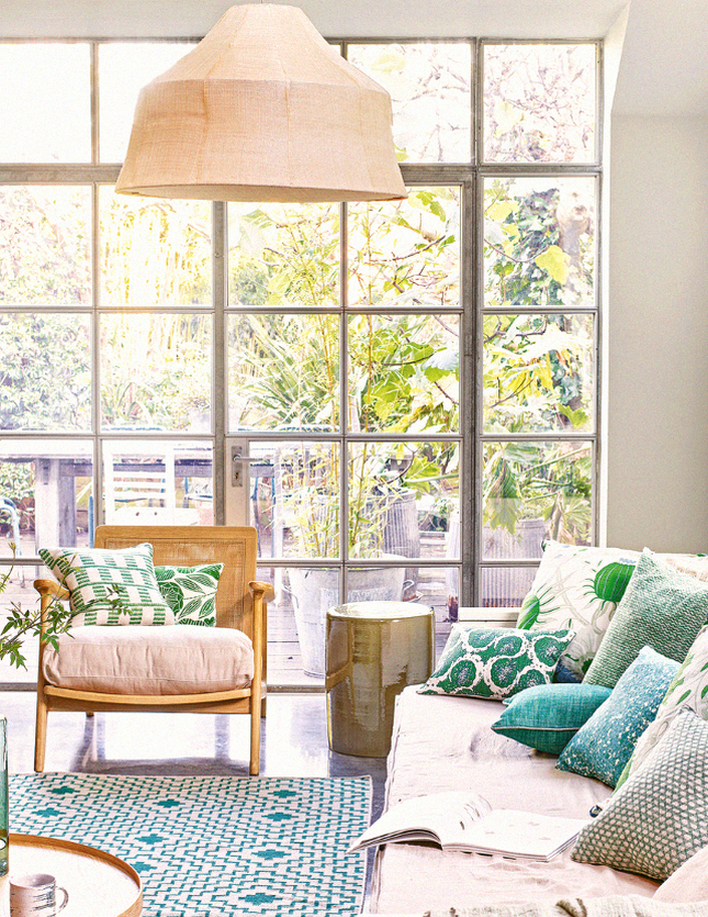 Sala de estar boho chic com sofá claro, poltrona de madeira e almofadas verdes próxima a janelas amplas