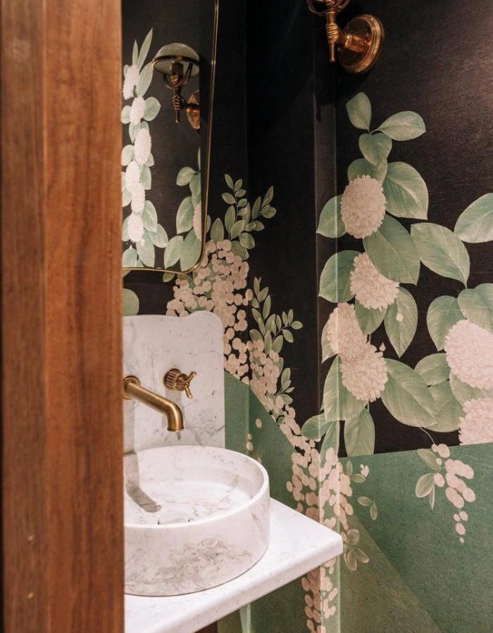 Banheiro pequeno preto com metais dourados e papel de parede floral verde com flores brancas.
