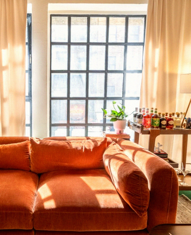 Sala contemporânea com sofá terracota em veludo, grandes janelas, cortinas claras, carrinho-bar e luz natural destacando a riqueza dos tons terrosos.